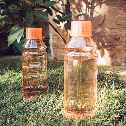 Two orange caped bottles, one is filled with a liquid, on grass with a tree on the background. The sun light casts soft shadows over the bottles. 