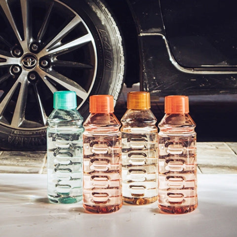 Four water bottles with a different colored caps ( green and orange ) arranged on a flat surface with car's tire in the background. the bottles are filled with liquids in different shades.