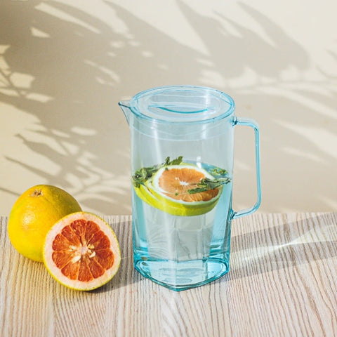 A blue acrylic jug is filled with water, lemon slices and amint leaves. the jug is placed next to a whole and a half lemon, with soft light casting shadows on the background.