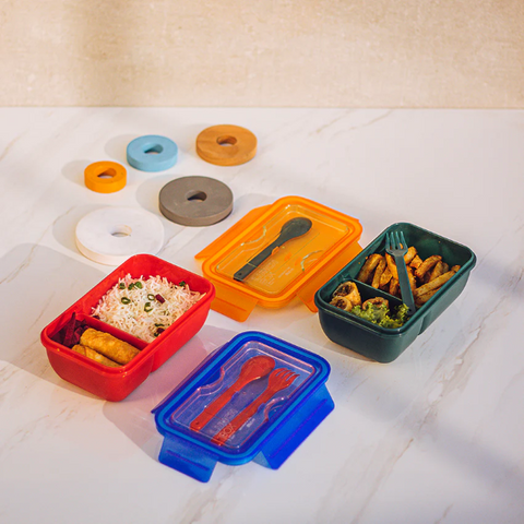 Colorful lunch boxes with food, including rice and vegetables, featuring different colors like red, green, and blue, with matching utensils inside.