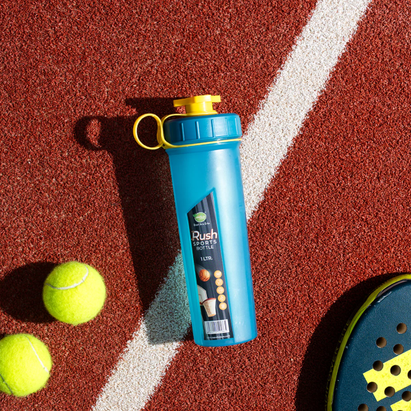 A blue rush sports bottle ( 1ltr ) with a yellow cap, placed on sports field. there are 2 tennis balls on the left side of bottle and a part of paddle on the right side. Highlighting a sporty, acticve lifestyle.