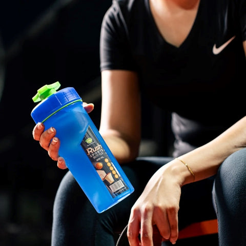 A person is holding a blue Rush Sports Bottle (800ml) with a green cap. the bottle is in the person's hand as he sits on bench, dressed in a athletic wear, with a dark, gym like background.