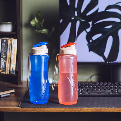 Two colour full water bottles, one in blue with a blue cap and the other is pink with an orange cap, both with a beige strap and placed on a wooden desk. A computer keyboard and monitor with a plant decor are visible  in the background.
