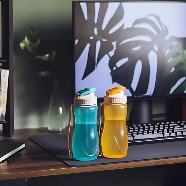 Two colordul water bottles, one turquoise and one yellow, both with a white cap and a beige carrying strap. they are placed on awooden desk, with a computer monitor displaying plant leaves in the background.