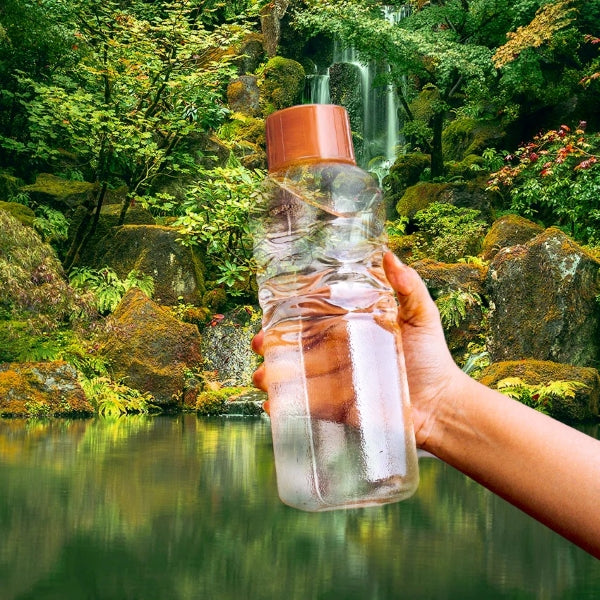A person holding a clear, plastic water bottle with an orange cap, standing against a serene natural backdrop of a waterfall and lush greenery